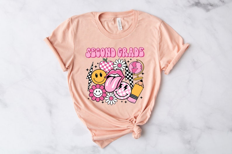 May include: A peach-colored t-shirt with the text "Second Grade" in pink letters. The shirt features a variety of pink and yellow graphic designs, including a smiley face, a flower, a checkered pattern, a tongue, a pencil, and a globe.