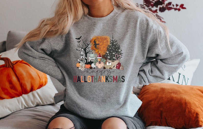 Puede incluir: Sudadera gris con un dise&ntilde;o gr&aacute;fico colorido que presenta un esqueleto, calabazas, un pavo, un &aacute;rbol de Navidad y el texto "Happy Hallothanksmas".