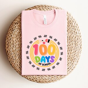 May include: A light pink t-shirt with the text "100 DAYS OF SCHOOL" in colorful bubble letters. The design includes a yellow circle with tally marks around the text. A pencil and apple are also included in the design.