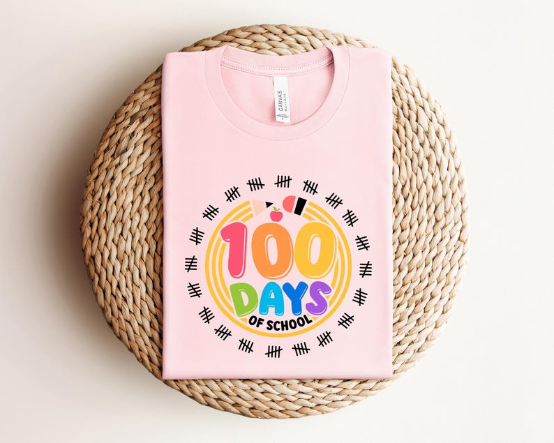 May include: A light pink t-shirt with the text "100 DAYS OF SCHOOL" in colorful bubble letters. The design includes a yellow circle with tally marks around the text. A pencil and apple are also included in the design.
