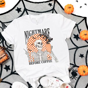 May include: White t-shirt with a black and white skeleton graphic holding a cup of coffee. The graphic is surrounded by a checkered pattern and bats. The text "Nightmare Before Coffee" is printed below the graphic.