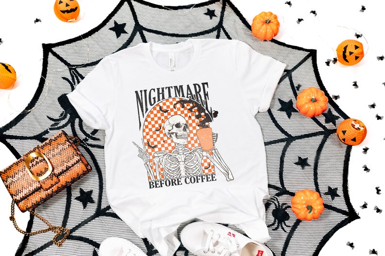 May include: White t-shirt with a black and white skeleton graphic holding a cup of coffee. The graphic is surrounded by a checkered pattern and bats. The text "Nightmare Before Coffee" is printed below the graphic.