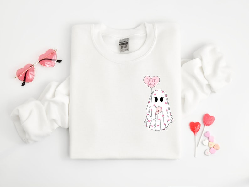 Op de afbeelding: Wit sweatshirt met een spook dat een hartvormige ballon vasthoudt met de tekst "Be My Boo". Het spook is bedekt met kleine roze hartjes.