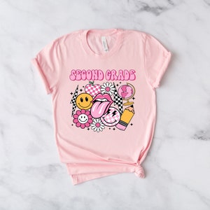 May include: Pink t-shirt with the text "Second Grade" in a retro font. The shirt features a graphic design of a smiley face, a tongue, a checkered pattern, a globe, a pencil, and flowers.