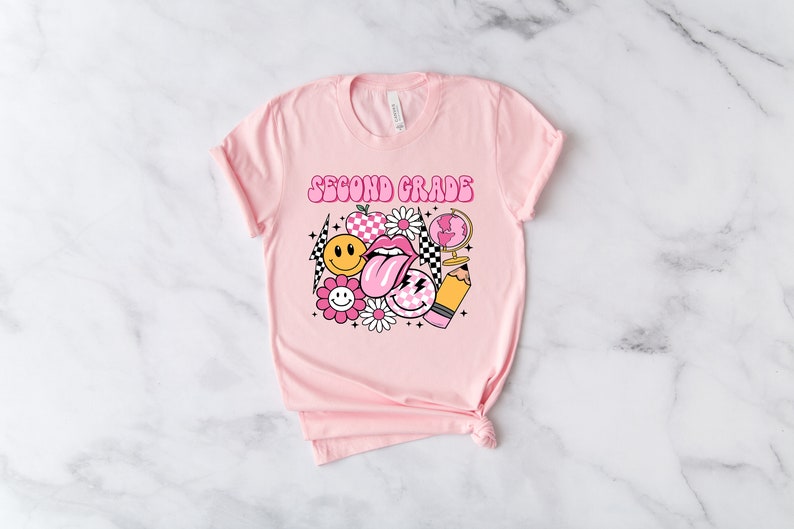 May include: Pink t-shirt with the text "Second Grade" in a retro font. The shirt features a graphic design of a smiley face, a tongue, a checkered pattern, a globe, a pencil, and flowers.
