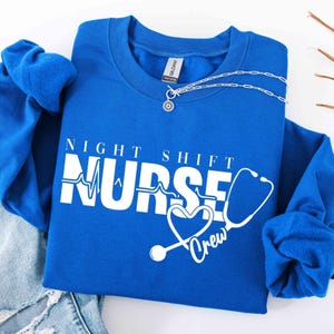 May include: Royal blue crewneck sweatshirt with the text "NIGHT SHIFT NURSE Crew" in white. The design includes a heartbeat line, a heart-shaped stethoscope, and a silver necklace.