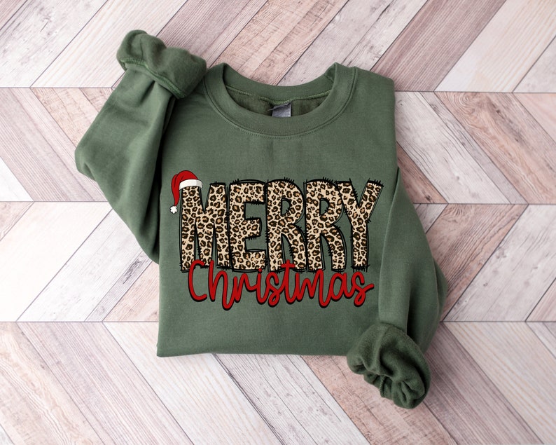 May include: A green sweatshirt with a leopard print "Merry Christmas" design. The word "Merry" has a red Santa hat on top.
