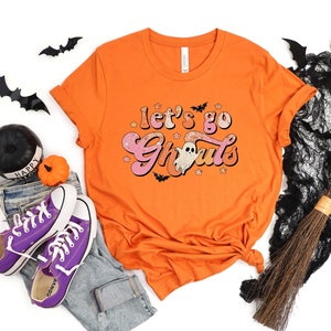 May include: Orange t-shirt with a pink and white graphic that says "Let's go Ghoul's". The graphic features a ghost and stars.