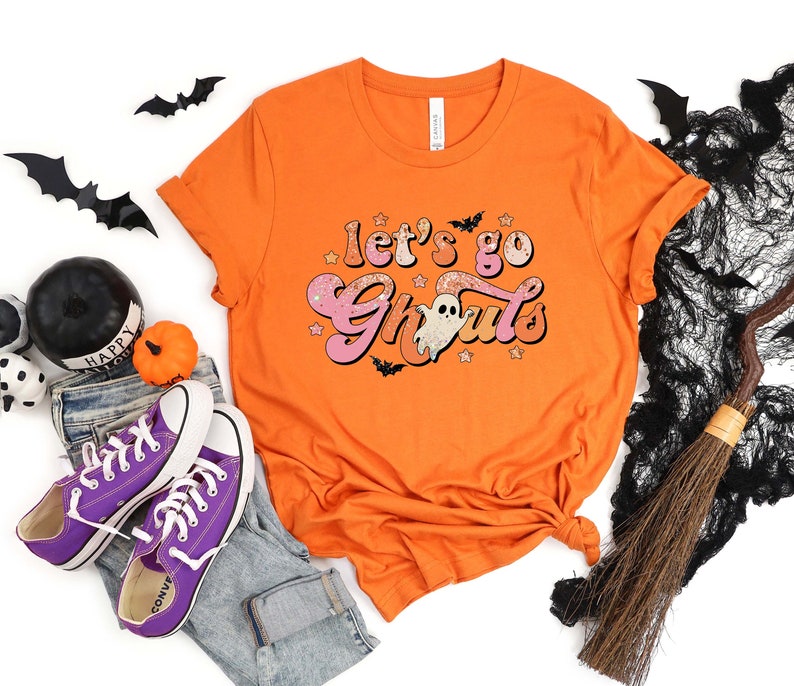 May include: Orange t-shirt with a pink and white graphic that says "Let's go Ghoul's". The graphic features a ghost and stars.