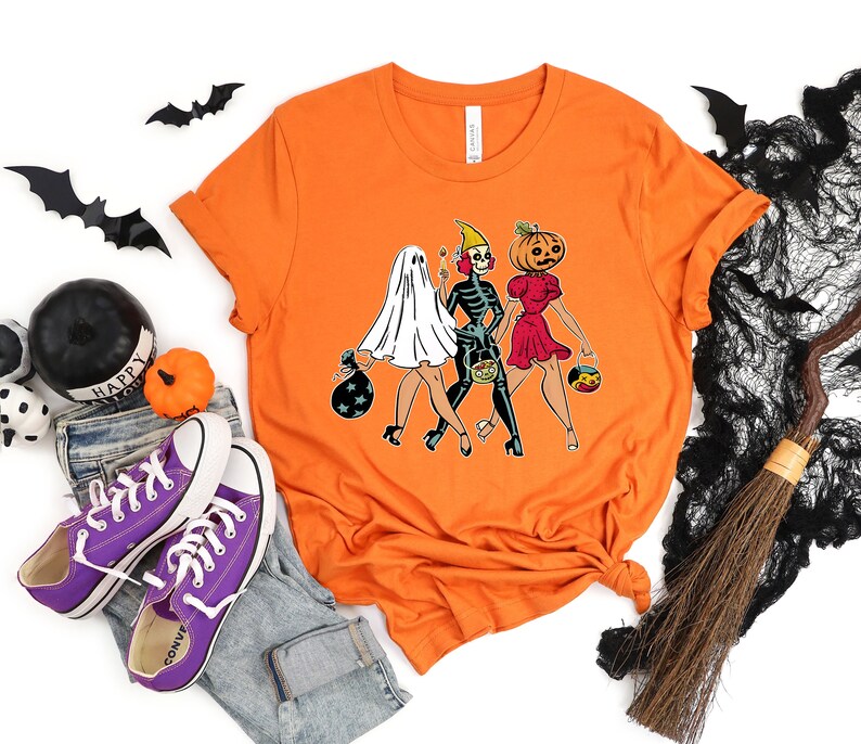 Pu&ograve; includere: Maglietta arancione con una grafica di tre donne vestite da fantasma, scheletro e zucca per Halloween. Le donne tengono caramelle e camminano insieme.