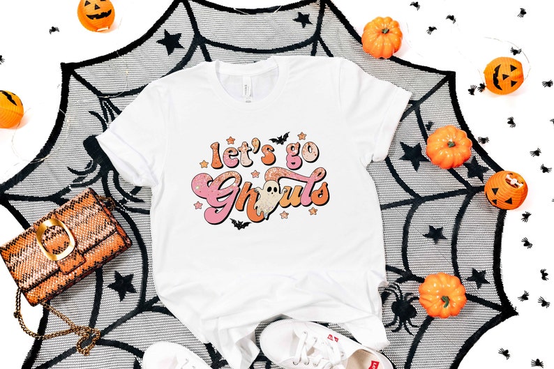 May include: White t-shirt with a pink and orange graphic that says "Let's go Ghouls" with a ghost in the middle. The graphic has a glittery effect.