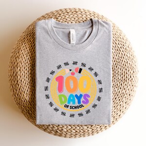 May include: A gray t-shirt with a colorful graphic celebrating 100 days of school. The design features the text "100 DAYS OF SCHOOL" in vibrant colors, surrounded by a circle of tally marks, pencils, and a yellow outline.