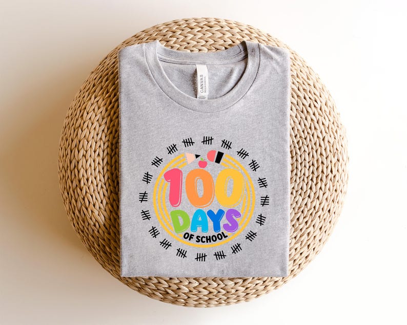 May include: A gray t-shirt with a colorful graphic celebrating 100 days of school. The design features the text "100 DAYS OF SCHOOL" in vibrant colors, surrounded by a circle of tally marks, pencils, and a yellow outline.