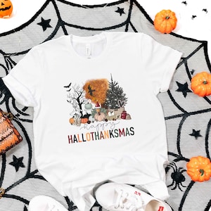 Puede incluir: Camiseta blanca con un dise&ntilde;o gr&aacute;fico de una escena tem&aacute;tica de Halloween y Acci&oacute;n de Gracias con el texto "Happy Hallothanksmas". El dise&ntilde;o incluye una calabaza, un &aacute;rbol de Navidad, un cuervo y un fantasma.