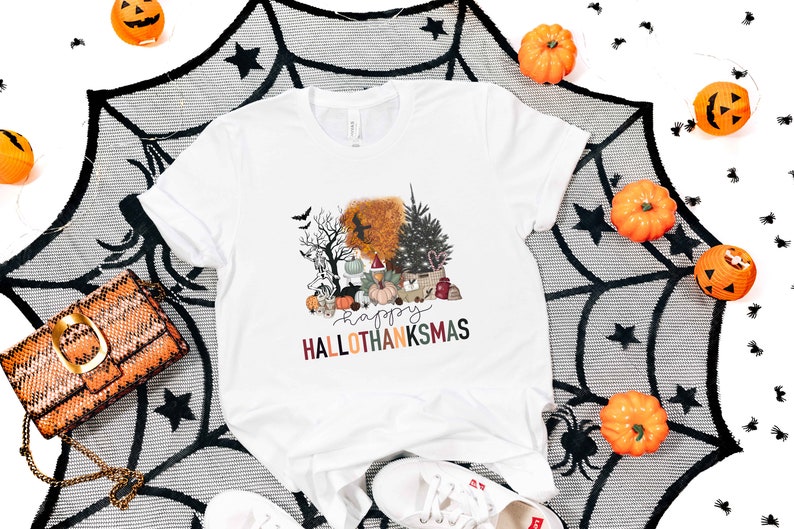 Puede incluir: Camiseta blanca con un dise&ntilde;o gr&aacute;fico de una escena tem&aacute;tica de Halloween y Acci&oacute;n de Gracias con el texto "Happy Hallothanksmas". El dise&ntilde;o incluye una calabaza, un &aacute;rbol de Navidad, un cuervo y un fantasma.