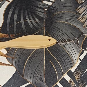 Peut inclure: Un porte-clés en bois en forme de poisson avec une chaîne et un anneau argentés. Le porte-clés est sur un fond à motifs noir et or.