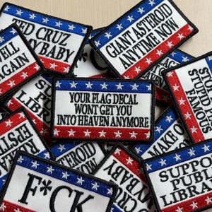 May include: A collection of rectangular embroidered patches featuring a red, white, and blue flag-inspired border with white stars. Each patch displays unique black text, including phrases like GIANT ASTEROID ANYTIME NOW, SUPPORT PUBLIC LIBRARIES, and YOUR FLAG DECAL WONT GET YOU INTO HEAVEN ANYMORE.