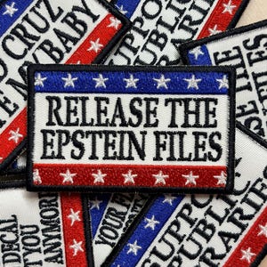 May include: A rectangular embroidered patch featuring a white background with a blue top stripe and a red bottom stripe, each containing white stars. The center text in black reads RELEASE THE EPSTEIN FILES, bordered by a thick black stitched edge.