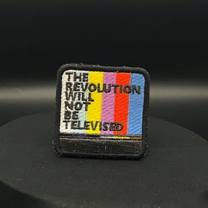 May include: Embroidered patch with the text "THE REVOLUTION WILL NOT BE TELEVISED" in white, set against a colorful background of vertical stripes. The stripes include yellow, red, purple, and blue. The patch has a black border.