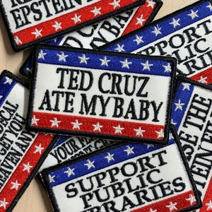 May include: A collection of rectangular embroidered patches featuring a red, white, and blue border with stars. The patches display various text including 'RELEASE THE EPSTEIN FILES', 'TED CRUZ ATE MY BABY', and 'SUPPORT PUBLIC LIBRARIES' in black.