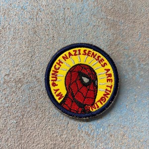 May include: A circular embroidered patch featuring a red and black masked character in the center against a yellow background with white radiating lines. The text MY PUNCH NAZI SENSES ARE TINGLING is stitched in red along the yellow border.