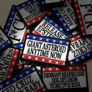 May include: A collection of rectangular embroidered patches featuring red, white, and blue borders with white stars. The patches display various black text slogans including GIANT ASTEROID ANYTIME NOW, F*CK, RELEASE EPSTEIN, and SUPPORT PUBLIC LIBRARIES.