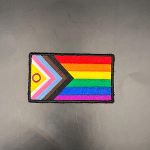 Pode incluir: Patch bordado com o design da bandeira Progress Pride. A bandeira inclui as cores tradicionais do arco-íris, juntamente com cores e símbolos adicionais que representam a inclusão. Borda preta.