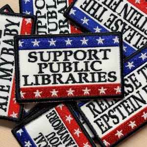 May include: A rectangular embroidered patch with a white center featuring the black text SUPPORT PUBLIC LIBRARIES. The top border is blue with white stars, and the bottom border is red with white stars. Other patches with various text are visible.
