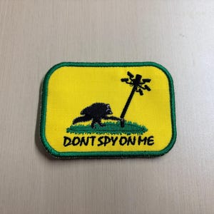 May include: A rectangular embroidered patch with a yellow background and a green border. It features a black silhouette of a creature pushing over a pole topped with surveillance cameras, with the text DONT SPY ON ME printed in black below.