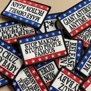 May include: A collection of rectangular embroidered patches featuring red, white, and blue borders with stars. The patches display various slogans in black text, including 'STOP MAKING STUPID PEOPLE FAMOUS', 'MAKE ORWELL FICTION AGAIN', and 'GIANT ASTEROID ANYTIME NOW'.