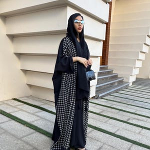 May include: A woman wearing a black abaya with a white and black geometric pattern. The abaya is paired with black pants, a black top, and a black head covering. She is also wearing sunglasses and carrying a small black purse. The background features a building with stairs.