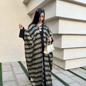 May include: A black and gold patterned abaya, a traditional garment, is worn over a black outfit. The abaya features a vertical gold stripe design. A small, gold handbag and gold-colored shoes complete the look. Sunglasses are on top of the head.