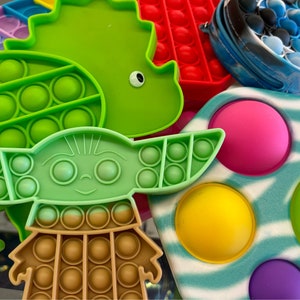 May include: A collection of colorful silicone fidget toys in various shapes, including a green dinosaur, a green turtle, and a Baby Yoda figure. Other shapes include a red rectangle and a blue tie-dye zippered pouch.