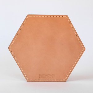 May include: A hexagonal leather coaster with stitched edges and the text "mothersproject goods" printed on the bottom.