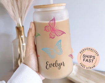 May include: A frosted glass tumbler with a bamboo lid and a straw. The tumbler has a brown background with colorful butterfly designs and the name "Evelyn" printed on the front. The tumbler is being held by a hand.