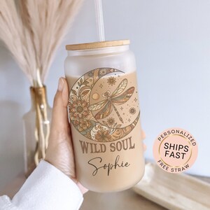 May include: A clear glass tumbler with a bamboo lid and a straw. The tumbler has a design of a crescent moon with a dragonfly and floral patterns. The text "Wild Soul Sophie" is printed on the tumbler.