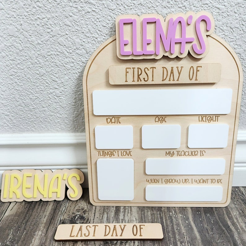 May include: Wooden sign with the text "ELENA'S FIRST DAY OF" and "IRENA'S LAST DAY OF" in pink and yellow letters. The sign has blank spaces for writing in the date, age, height, things they love, their teacher's name, and what they want to be when they grow up.