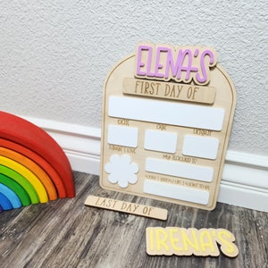 May include: Wooden sign with the text "ELENA'S FIRST DAY OF" and "LAST DAY OF" with blank spaces to fill in information about the first and last day of school. The sign also has spaces to write the date, age, favorite things, teacher's name, and what the child wants to be when they grow up. The sign is decorated with a pink flower and the name "IRENA'S" is written in yellow on a separate wooden sign.