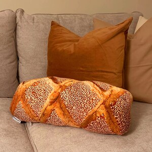 May include: A brown and tan pillow shaped like a loaf of bread with sesame seeds on top. The pillow is sitting on a light gray couch with two other pillows behind it.