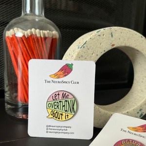 Può includere: Una spilla smaltata colorata con la scritta "Let Me Overthink About It" in un design a fumetto. La spilla è su una carta bianca con il logo "The NeuroSpicy Club". Un barattolo di vetro di fiammiferi e una pietra decorativa sono sullo sfondo.