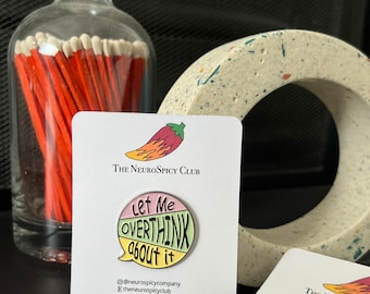 May include: A colorful enamel pin with the text "Let Me Overthink About It" in a speech bubble design. The pin is on a white card with "The NeuroSpicy Club" logo. A glass bottle of matches and a decorative stone are in the background.