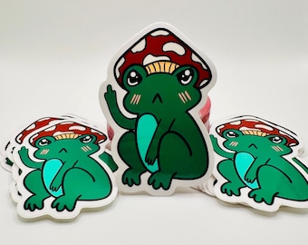 Puede incluir: Un conjunto de pegatinas de ranas verdes con sombreros de setas rojas y blancas. Las ranas están sentadas con los brazos cruzados y tienen una expresión de enfado en sus caras.