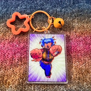 May include: A clear acrylic keychain with a cartoon illustration of a superheroine with blue hair and the word "feed" on one fist. The keychain has an orange star-shaped charm, a round ring, and a bell.