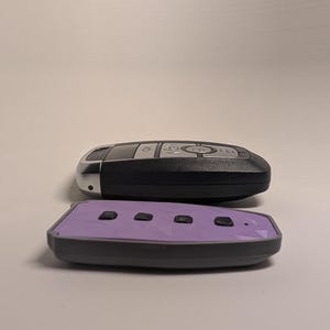 May include: A black car key fob with silver buttons positioned behind a separate light purple key fob cover featuring four black buttons and a geometric pattern on its surface, both resting on a plain, neutral-colored surface.