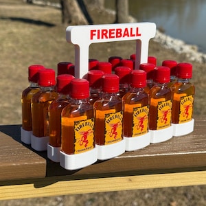 Może przedstawiać: Biały plastikowy pojemnik z uchwytem, oznaczony napisem "FIREBALL", zawiera 12 małych butelek Fireball Cinnamon Whisky. Butelki mają czerwone nakrętki i są ułożone w siatkę.