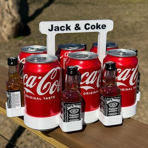 Może przedstawiać: Biały plastikowy sześciopak z uchwytem, oznaczony jako "Jack & Coke". Zawiera sześć puszek Coca-Coli i dwie mini butelki Jack Daniel's Tennessee Whiskey.