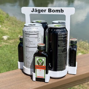 Puede incluir: Un soporte de plástico blanco para Jäger Bomb contiene cuatro latas y dos botellas pequeñas. El soporte tiene la inscripción "Jäger Bomb". Las latas son plateadas y negras, y las botellas son de color marrón oscuro con etiquetas verdes.