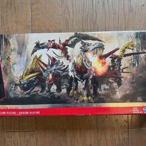 May include: A rectangular box featuring Dinobots from the Transformers franchise. The box art depicts several robotic dinosaurs in action, with a fiery explosion. The box has red and black accents and the Transformers logo.