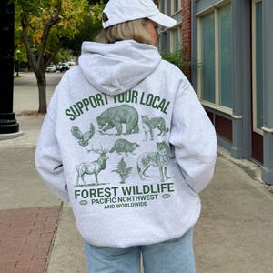 Puede incluir: Una sudadera gris con un estampado verde que dice "Support Your Local Forest Wildlife Pacific Northwest and Worldwide". El estampado presenta ilustraciones de varios animales, incluyendo un oso, un b&uacute;ho, un lobo, un ciervo y un mapache.
