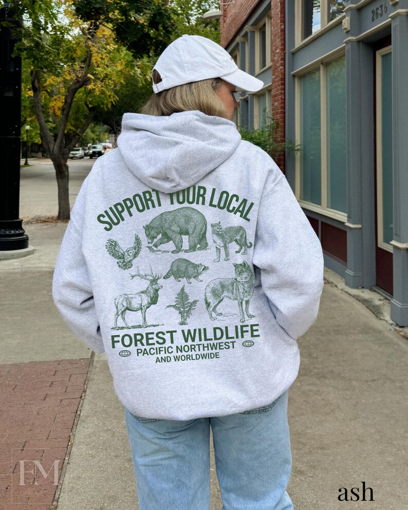 Puede incluir: Una sudadera gris con un estampado verde que dice "Support Your Local Forest Wildlife Pacific Northwest and Worldwide". El estampado presenta ilustraciones de varios animales, incluyendo un oso, un b&uacute;ho, un lobo, un ciervo y un mapache.
